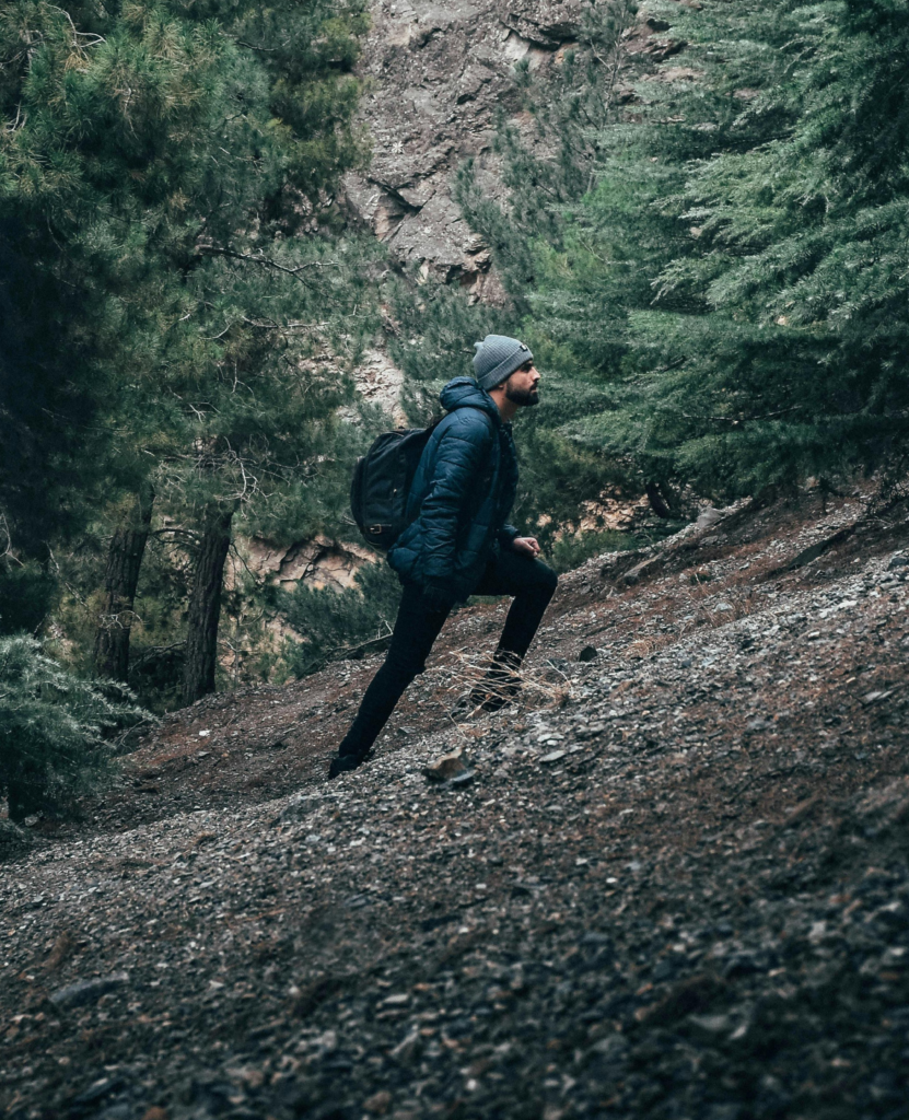 profile view of a man wearing a grey beanie, puffer jacket and black rucksack backpack walking up a steep mountain